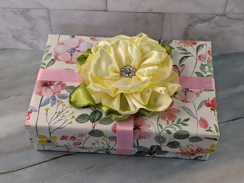 A pre-wrapped gift box with floral wrapping paper, a pink satin ribbon and a handmade yellow satin flower gift topper that has a beautiful gem button center.