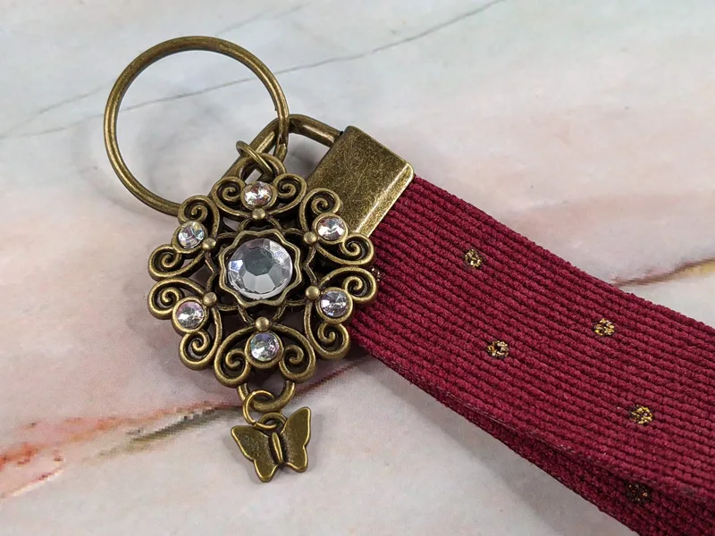 A wristlet keyring made of burgundy corduroy fabric with a golden pendant that has rhinestones and a butterfly charm.