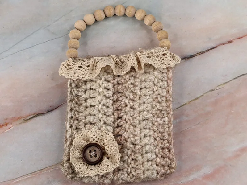 A tan gift card holder crocheted with tan yarn, handmade with cream lace and a wooden beaded handle.
