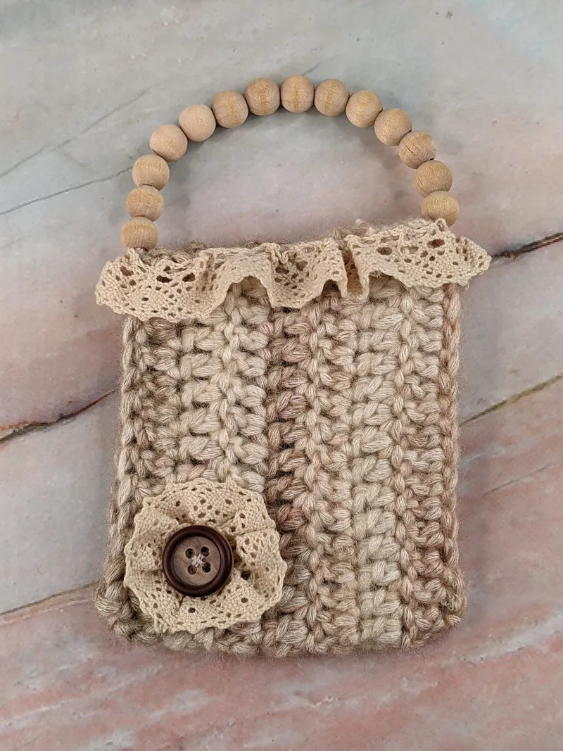 A crocheted gift card holder made with tan yarn, cream lace, and has natural wooden beads for a handle.