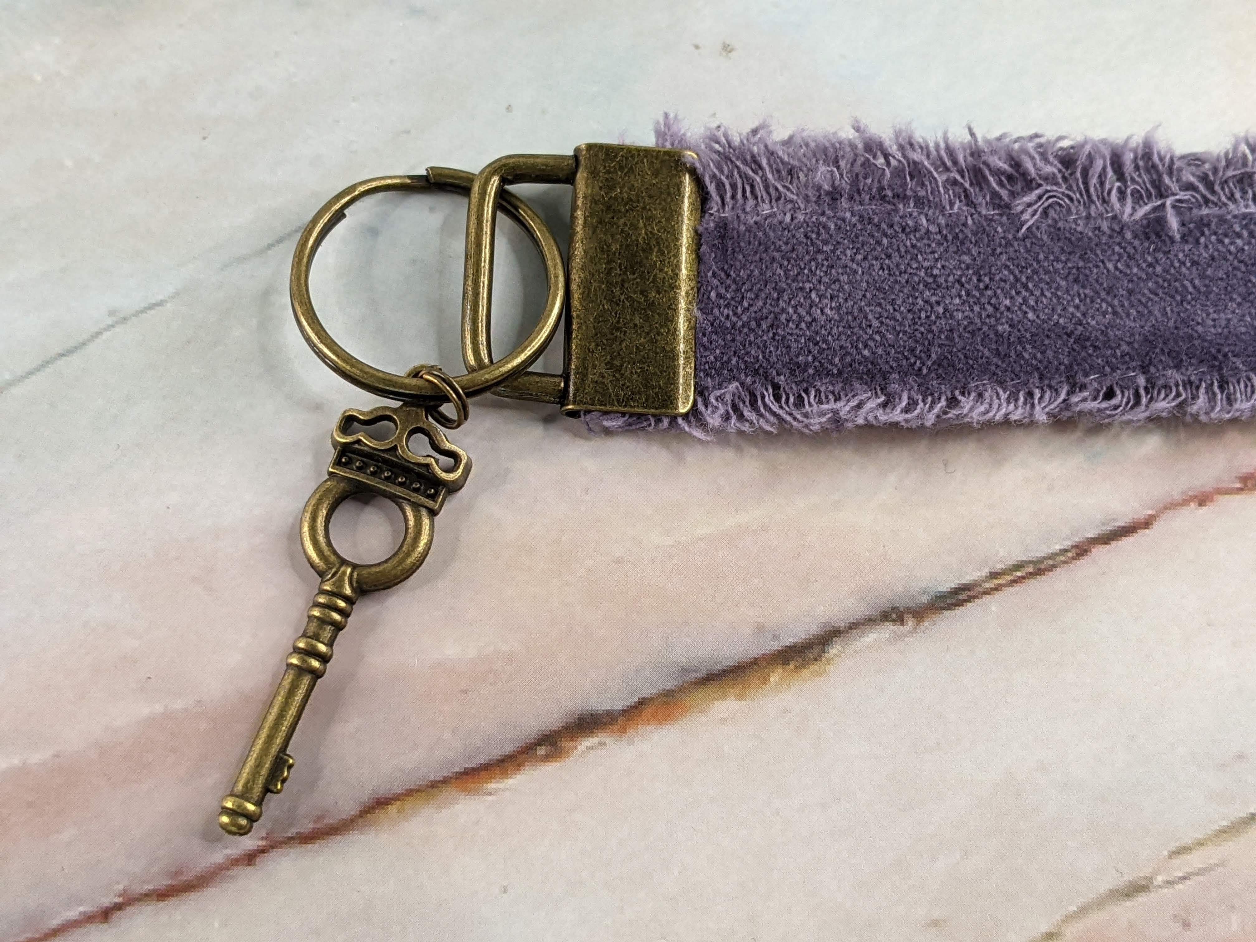 A dusty-purple fabric wristlet keychain with a golden metal key charm on a keyring. 