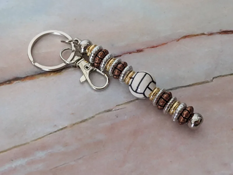 A bag charm keychain that has silver, gold, and bronze toned beads along with a white bead that looks like a volleyball.