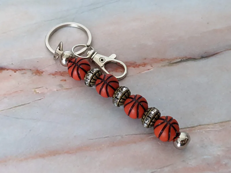 A beaded bag charm with a silver metal keyring, rhinestone accented golden metal beads and 4 orange basketball shaped beads.