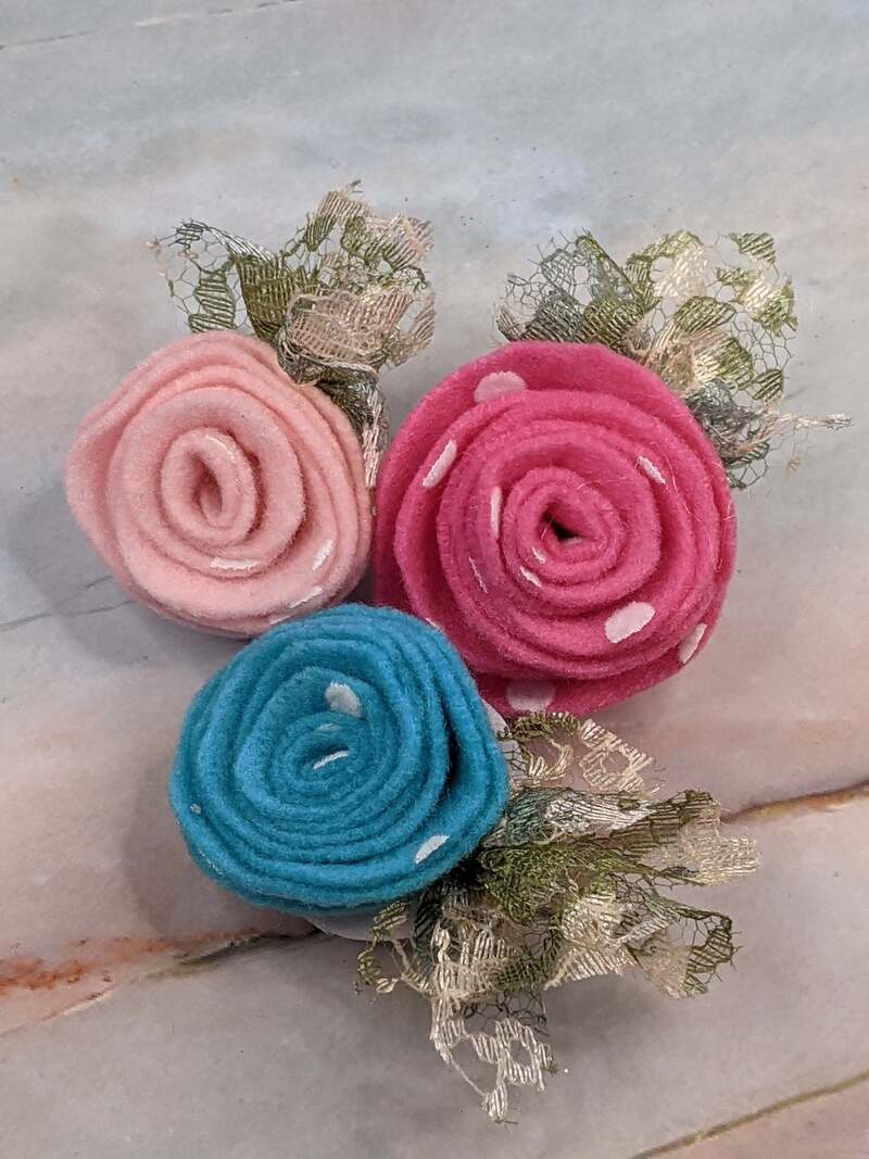 A set of three felt fabric handmade rosettes in light pink, dark pink and aqua blue, designed to clip onto a wrapped present.