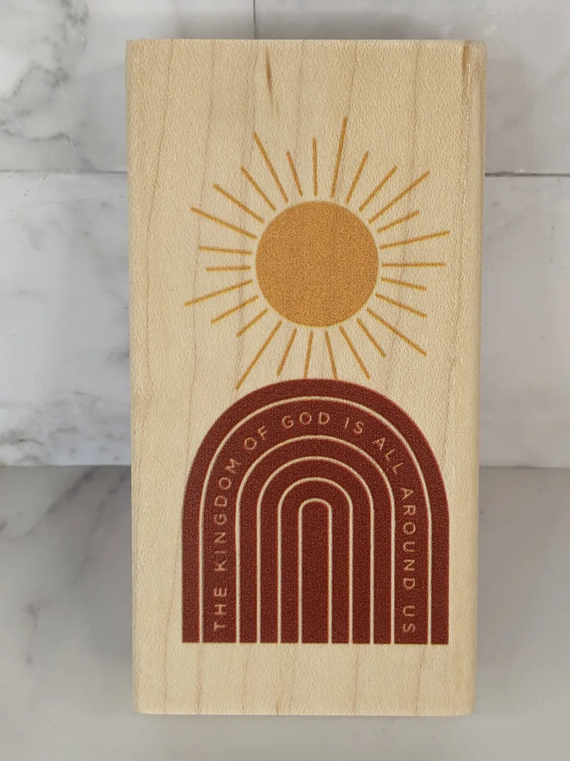 A wood mounted rubber stamp with an image of a sunshine over a rainbow shape with the words "The Kingdom of God is all around us."